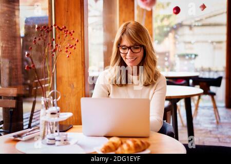 Confident mature woman in smart casual wear working on laptop while sitting near window in cafe. Small business. Stock Photo