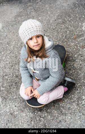 Adorable little girl in winter wool clothes sitting on skateboard outdoors in backyard - Smiling little girl playing with skateboard - Playful childho Stock Photo