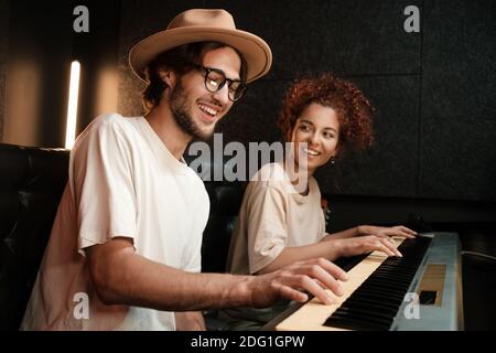 Young stylish musicians happily playing on piano keyboard recording song in modern recording studio Stock Photo