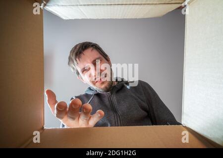Young disappointed and displeased man is opening gift and looking inside cardboard box. Stock Photo