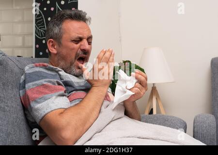 Unwell adult man sneezing on sofa holding napkin or handkerchief in hand as cold flu season symptoms sars covid pandemic concept Stock Photo