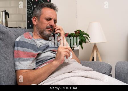 Sick adult man making wait busy stay gesture using index finger talking on phone resting on sofa at home having sars influenza flu covid19 cold sympto Stock Photo