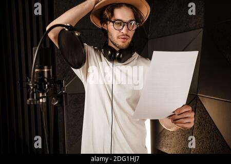 Young thoughtful male singer intently reading text of new song in sound recording studio Stock Photo