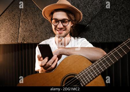 Young attractive man with guitar happily using smartphone in modern sound recording studio Stock Photo