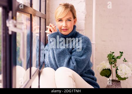 Portrait of beautiful blond haired woman wearing turtleneck sweater while relaxing at the window at home. Stock Photo