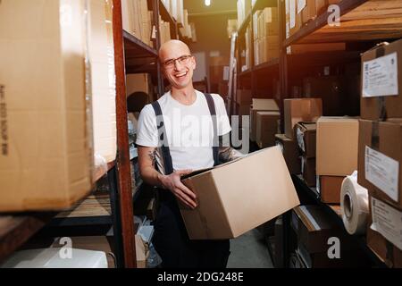Emotional warehouse worker carrying box, standing between tall rows of shelves Stock Photo