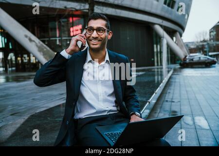Smiling mixed race businessman chatting on mobile device holding laptop Stock Photo