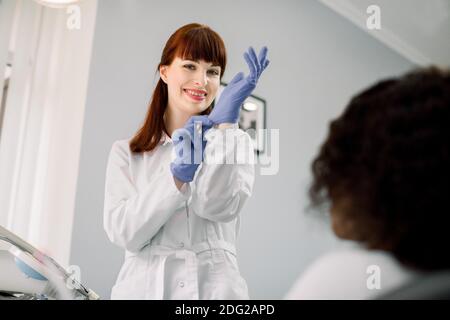 Beautiful Caucasian female dentist puts on blue rubber gloves, standing in a dental office, ready for the examination and treatment of her female Stock Photo