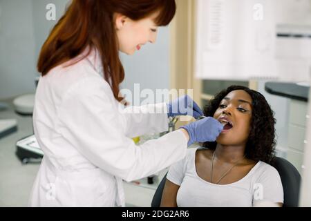 Young African female patient at dental office, opening mouth while smiling woman dentist in uniform and blue latex gloves, checking condition of teeth Stock Photo