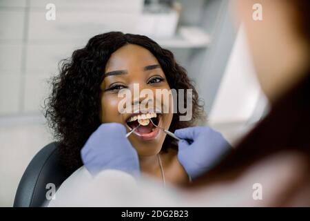 Close up of young beautiful smiling afro lady with beautiful white teeth, sitting in dental chair, while female dentist cures her teeth. Dentist Stock Photo