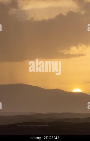 Mountainous landscape at sunset with a distant curtain of rain on the horizon and the sun disk hiding Stock Photo