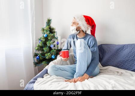 Covid-19 and pandemic New Normal concept. Young man wearing face mask and Santa hat sitting on couch, holding red mug of coffee, alone, in front of Stock Photo