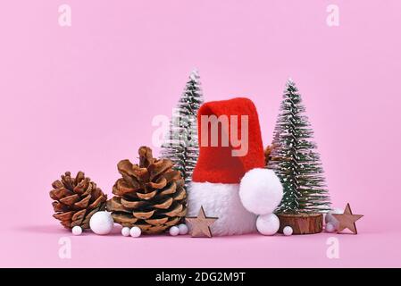 Christmas arrangement with red Santa hat, pine cone, star ornaments and small Christmas trees on pink background Stock Photo