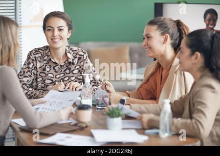 Multi-ethnic group of smiling businesswomen discussing project during meeting in office with remote participant in background, copy space Stock Photo