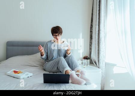 Emotional woman in glasses, casual home clothes having video chat on her laptop sitting on bed and drinking coffee. Remote working from home. Time at Stock Photo