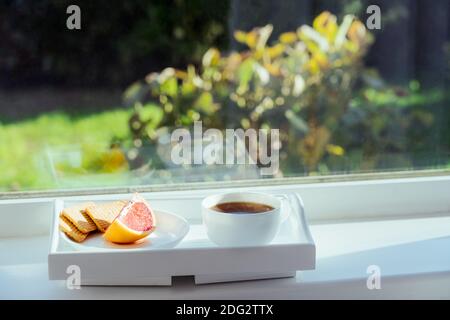 Cup of hot morning coffee, tea, grapefruit slice and cookies on the tray staying on the windowsill, indoors with green sunny garden background. Home Stock Photo