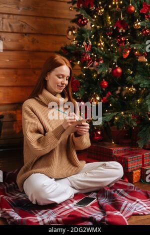 Cheerful dreamy young woman writing wish list for new year on background of Christmas tree Stock Photo