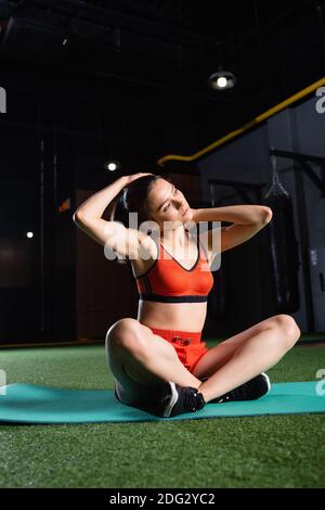 young sportswoman sitting on fitness mat and warming up neck in gym Stock Photo