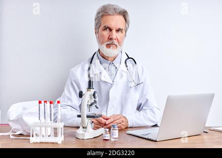 portrait of confident bearded doctor man in laboratory, doctor in white robe sits looking at camera, using laptop Stock Photo