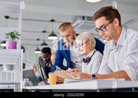 manager male is working in the office, caucasian guy looking at paper concentrated, thinking, while others are coworking together, focus on man Stock Photo