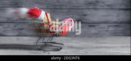 Fast small shopping cart with presents, christmas decoration, sweets and Santa Claus hat is driving on rustic wooden planks for last minute online gif Stock Photo