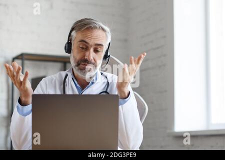 Portrait of senior grey-haired male doctor in his office using laptop for video chat with a patient. Online consultation with doctor for diagnoses and Stock Photo