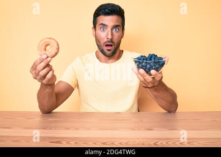 Young hispanic man eating breakfast holding donut and blueberries in shock face, looking skeptical and sarcastic, surprised with open mouth Stock Photo