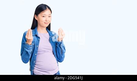 Young beautiful chinese woman pregnant expecting baby doing money gesture with hands, asking for salary payment, millionaire business Stock Photo