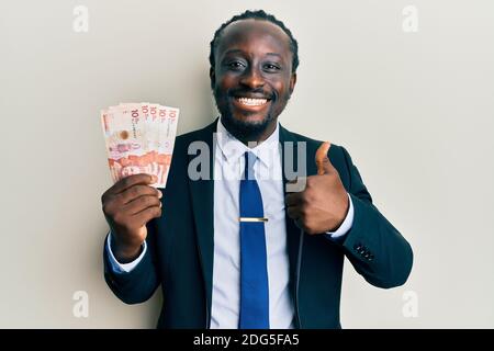 Handsome young black man wearing business suit holding 10 colombian pesos smiling happy and positive, thumb up doing excellent and approval sign Stock Photo