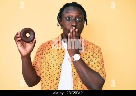 Young african american man with braids holding donut covering mouth with hand, shocked and afraid for mistake. surprised expression Stock Photo