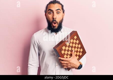 Young arab man holding chess scared and amazed with open mouth for surprise, disbelief face Stock Photo