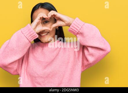Young asian woman wearing casual winter sweater doing heart shape with hand and fingers smiling looking through sign Stock Photo