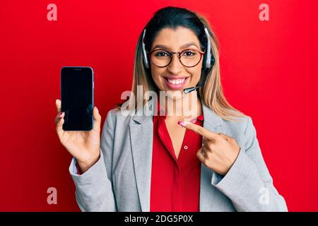 Beautiful brunette woman wearing operator headset showing smartphone screen smiling happy pointing with hand and finger Stock Photo