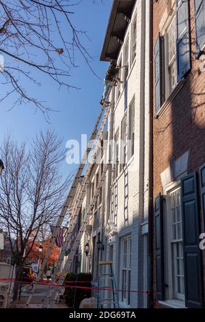 Alexandria, VA, USA 11-28-2020: A house  painter is on top of an industrial size aluminum extension ladder painting the exterior surface of an old bri Stock Photo