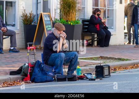 Alexandria, VA, USA 11-28-2020: A white bearded elderly street musician wearing boots, jeans and flat hat sitting on stool by the sidewalk is playing