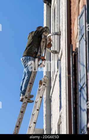 A hispanic house painter with paint stained work clothes is on top of a steel construction ladder painting the exterior surface of an old brick house. Stock Photo