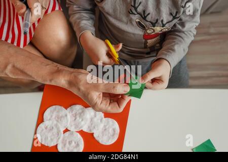 A Little boy with grandmother make Christmas craft, greeting card. Top view boy cuts a triangle out of paper with scissors. Stock Photo