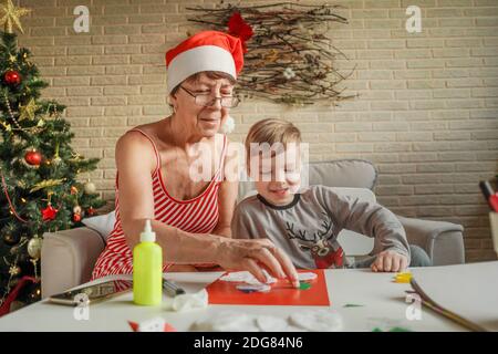 A Little boy with grandmother make Christmas craft, greeting card. Christmas tree and decoration on background. Stock Photo