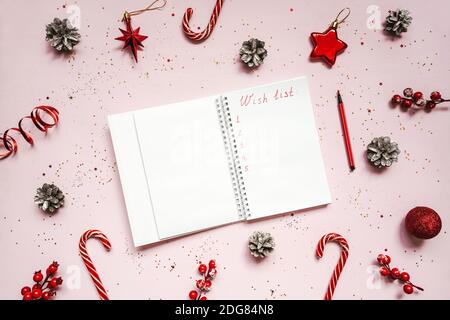 Wish list of presents text on notebook, christmas concept. Red baubles and pine cones on pink background. Stock Photo
