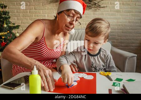 A Little boy with grandmother make Christmas craft, greeting card. Christmas tree and decoration on background. Stock Photo