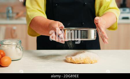 Senior woman with black apron sifting flour on dough using metallic sieve Happy elderly chef with bonete preparing raw ingredients to baking traditional bread sprinkling, sieving in the kitchen. Stock Photo
