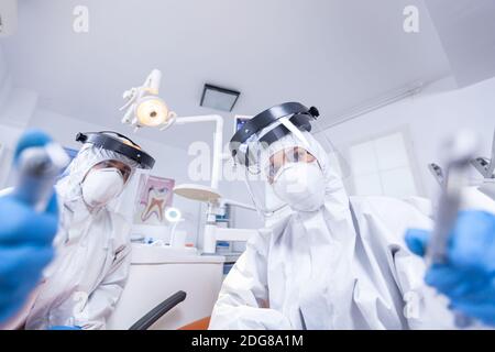 Patient pov of orthodontist and assistan doing procedure for mouth hygine in coverall. Stomatology team wearing safety equipment against coronavirus treating patient. Stock Photo