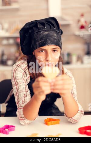 Grandchild looking at heart shaped pastry on christmas day wearing apron and bonette. Happy cheerful joyfull teenage girl helping senior woman preparing sweet cookies to celebrate winter holidays. Stock Photo