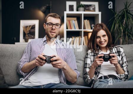 Joyful young couple sitting at home and playing video games together. Beautiful woman and handsome man relaxing on grey couch and using joysticks for Stock Photo