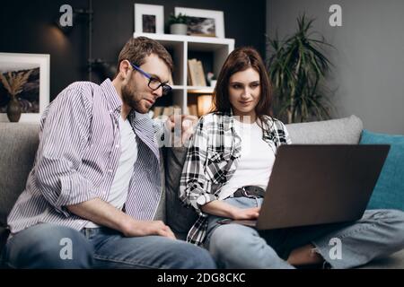 Young man and woman relaxing on comfortable sofa and talking during video call on laptop. Concept of people and technology. Stock Photo