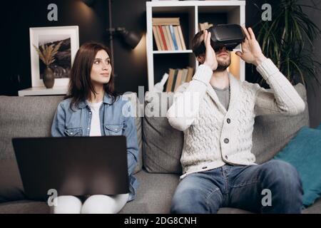 Attractive man and woman relaxing together at home and using VR headset with modern laptop. Young couple playing virtual games while sitting on couch. Stock Photo