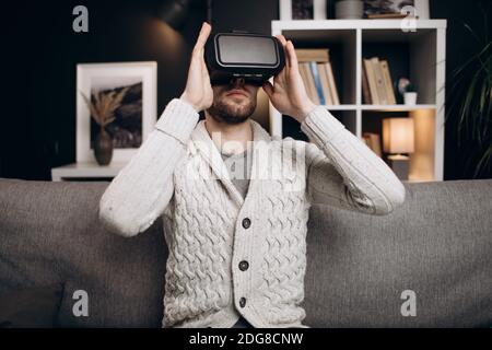 Handsome man wearing VR headset at home while relaxing on grey couch at home. Concept of modern device and innovation. Stock Photo