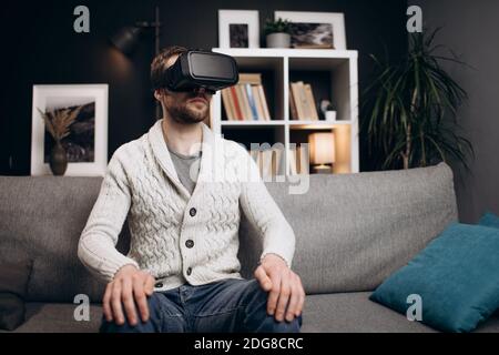 Young guy in casual clothing using modern VR headset during leisure time at home. Handsome man sitting on couch and using modern technology. Stock Photo