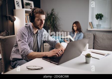 Handsome man in headset sitting at table and working on portable laptop. Young lady sitting on background and reading interesting book. Concept of dom Stock Photo