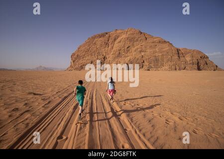 Young brother and sister are running through the Wadi Rum desert following the car tracks that leads to the huge rock mountain place of the Lawrence o Stock Photo